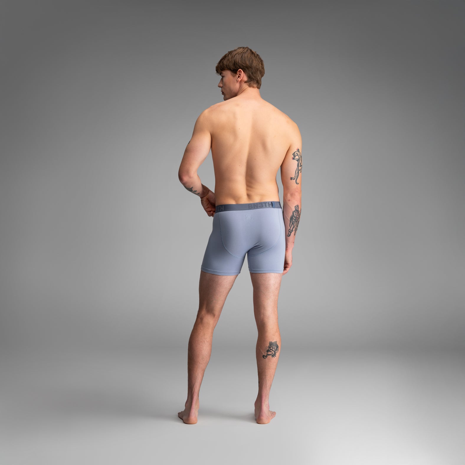 A man stands with his back to the camera wearing light blue boxer briefs against a smooth gray studio background. BN3TH Essentials Boxer Brief: Dusty Blue