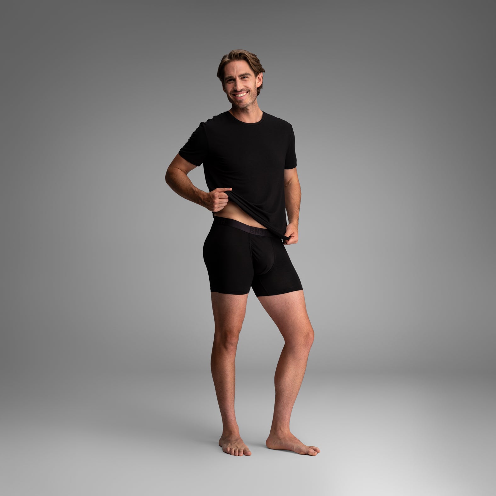 A man modeling a black short-sleeve t-shirt and matching black boxer briefs against a neutral gray studio background. . Essentials Boxer Brief: Black