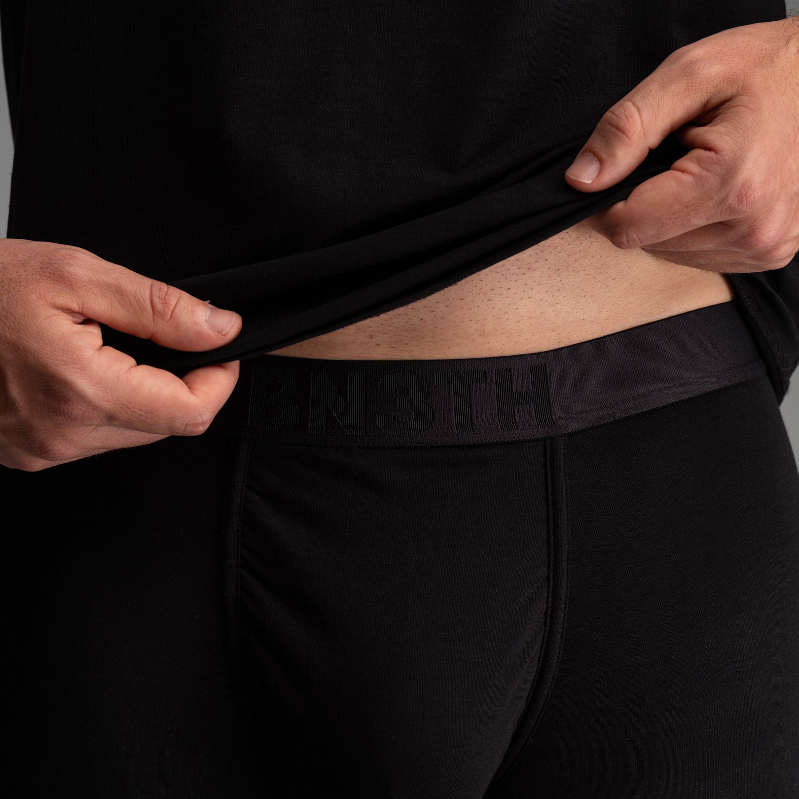 Close-up of a person wearing black underwear and lifting the hem of a black shirt to show the black branded waistband with the word 'BN3TH' embossed.