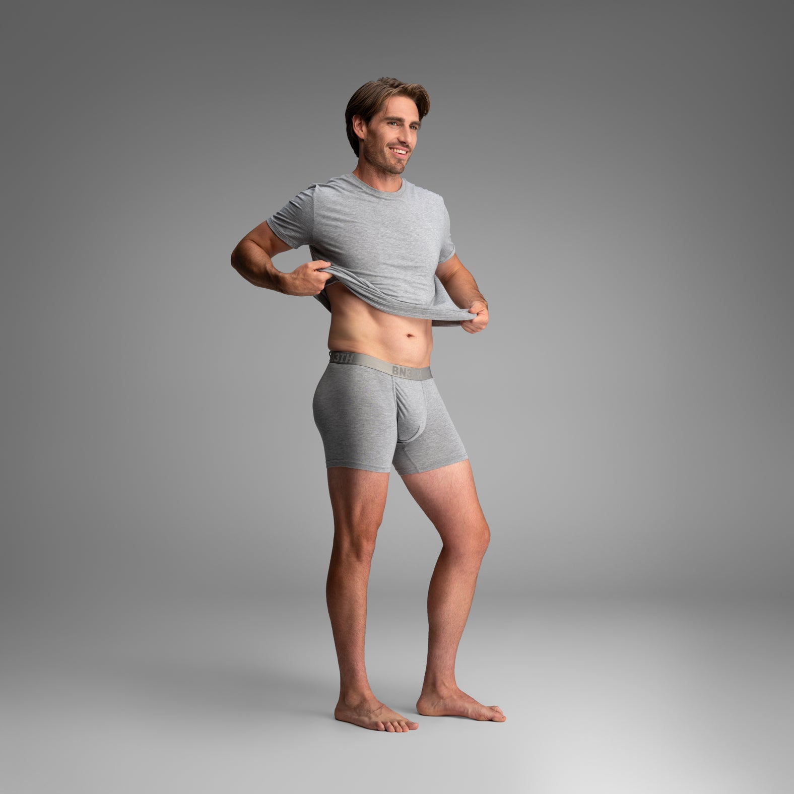 A man modeling a light gray crewneck t-shirt and matching gray boxer briefs, lifting the shirt to show fit against a neutral gray studio background.
