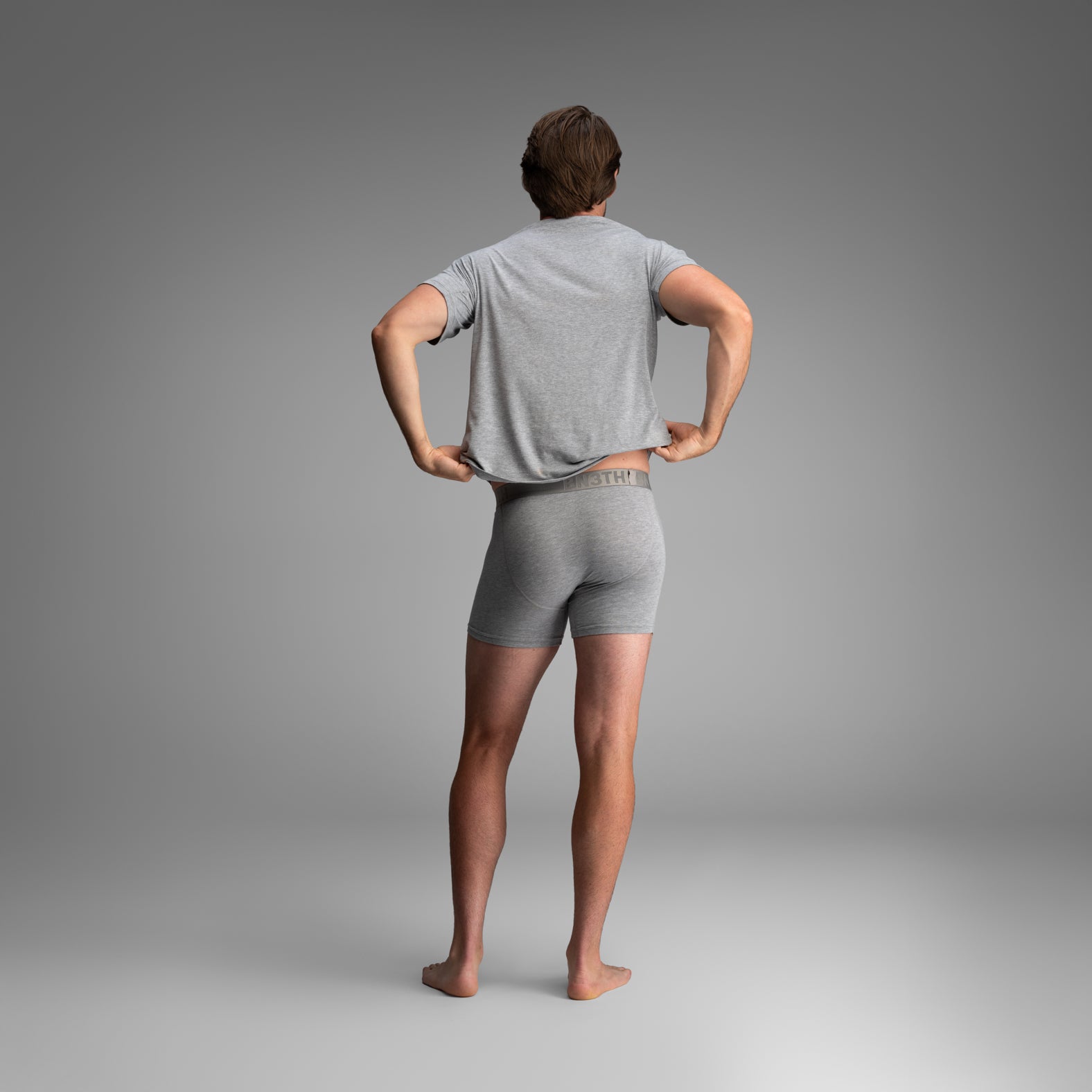 A man shown from behind wearing a light gray t-shirt and matching gray boxer briefs, standing against a neutral gray studio background. Essentials Boxer Brief: Heather Grey